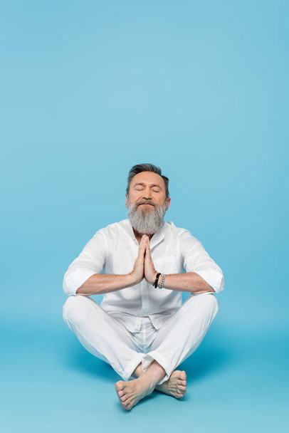 bärtiger Mann mit geschlossenen Augen und betenden Händen meditiert in lockerer Pose auf blauem Grund  - Foto, Bild