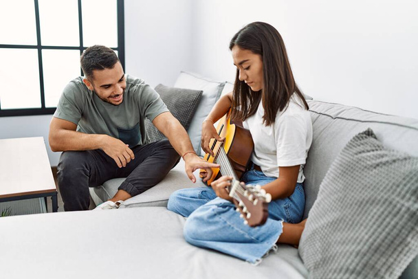 Joven pareja latina tocando la guitarra clásica sentada en el sofá en casa. - Foto, imagen