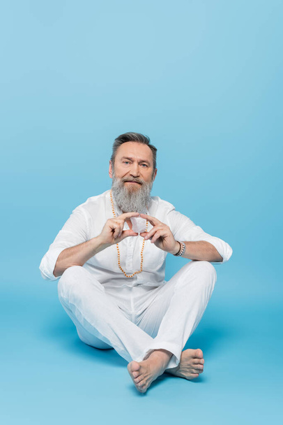 guru tutor sênior mostrando hakini mudra enquanto sentado com pernas cruzadas em azul  - Foto, Imagem