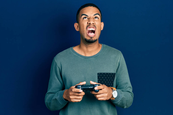 Νεαρός Αφροαμερικάνος που παίζει βιντεοπαιχνίδι κρατώντας το χειριστήριο θυμωμένος και τρελός ουρλιάζοντας απογοητευμένος και έξαλλος, φωνάζοντας με θυμό κοιτάζοντας ψηλά.  - Φωτογραφία, εικόνα