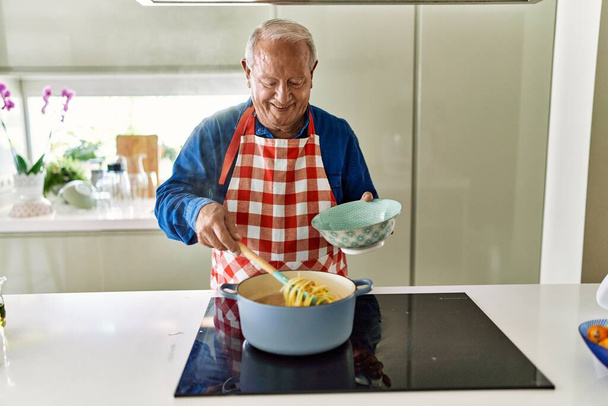 Hombre mayor sonriendo confiado cocinando espaguetis en la cocina - Foto, Imagen