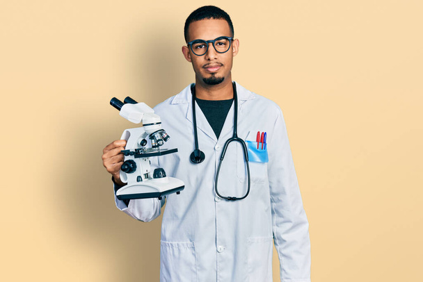 Genç Afrikalı Amerikalı adam, bilim adamı üniforması giyiyor. Mikroskop tutuyor. Düşünme tavrı takınıyor.  - Fotoğraf, Görsel