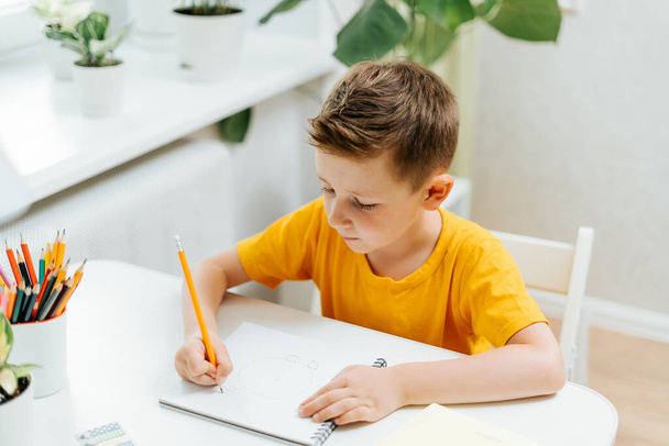 Дистанционное обучение онлайн. Белый улыбчивый мальчик учится дома с книгой, пишет в блокноте и делает домашнее задание. Мыслящий ребенок сидит за столом с блокнотом. Возвращение в школу. - Фото, изображение