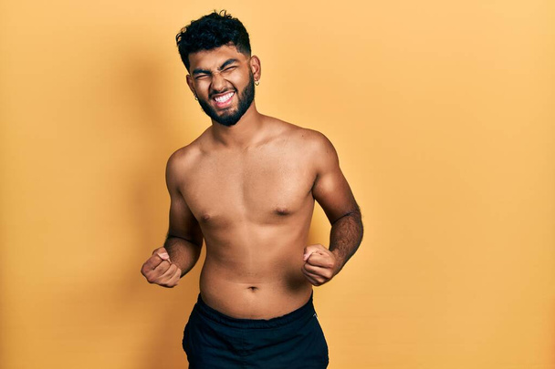 Arabischer Mann mit Bart in Badebekleidung ohne Hemd sehr glücklich und aufgeregt und macht Siegergeste mit erhobenen Armen, lächelnd und schreiend nach Erfolg. Festkonzept.  - Foto, Bild