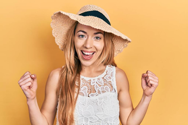 Jeune femme caucasienne portant un chapeau d'été célébrant surpris et étonnés pour le succès avec les bras levés et les yeux ouverts. concept gagnant.  - Photo, image