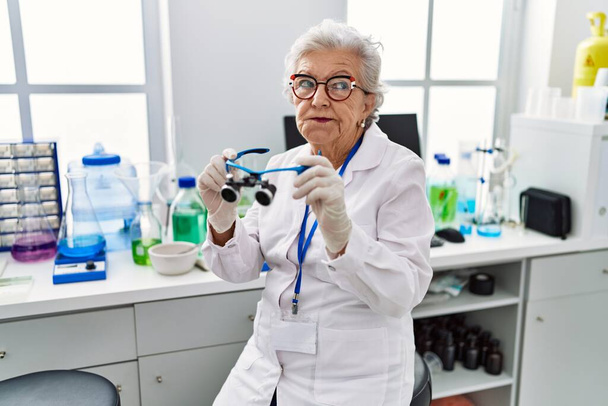 Старшая женщина с седыми волосами работает в научной лаборатории, используя увеличительные очки, улыбаясь, глядя в сторону и думая:.  - Фото, изображение