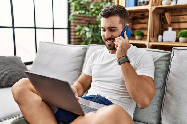 Genç İspanyol adam dizüstü bilgisayar kullanıyor ve evde akıllı telefonla konuşuyor. - Fotoğraf, Görsel