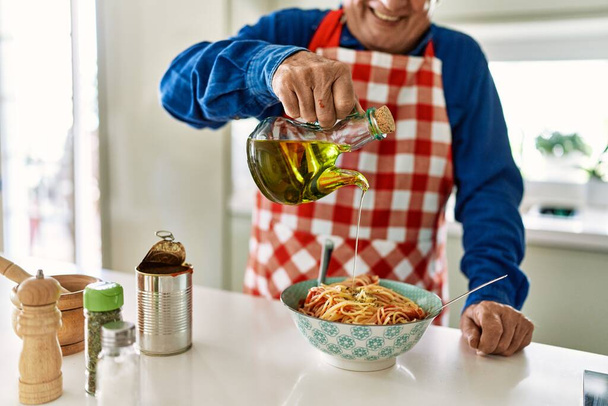 Hombre mayor sonriendo confiado vertiendo aceite en espaguetis en la cocina - Foto, imagen