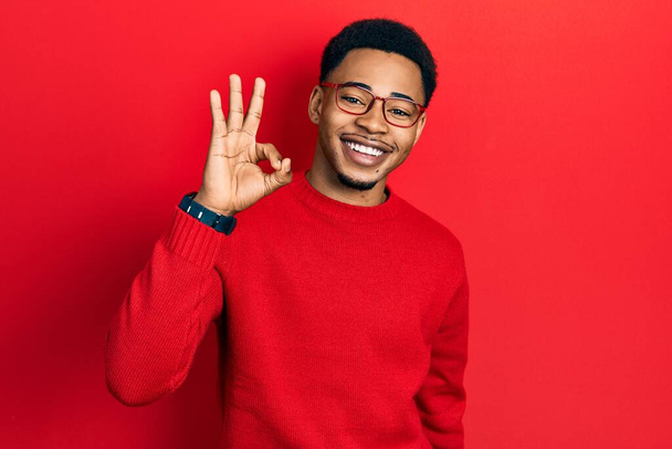 Jeune homme afro-américain portant des vêtements décontractés et des lunettes souriant positif faire ok signe avec la main et les doigts. expression réussie.  - Photo, image