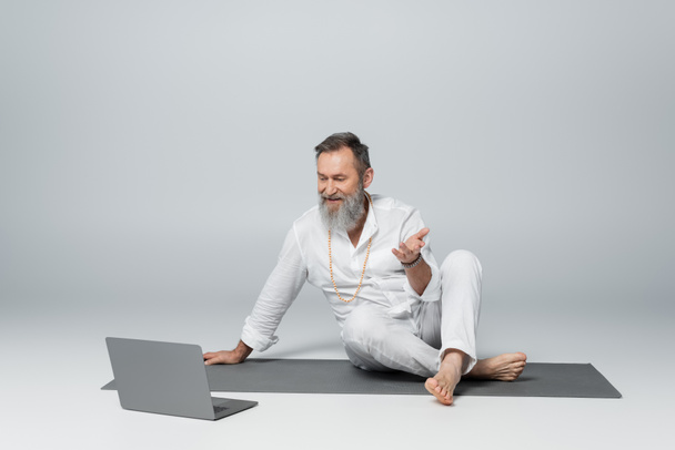 mentor gurú sentado en esterilla de yoga con las piernas cruzadas y hablando cerca del ordenador portátil en gris - Foto, Imagen