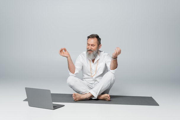 maestro de yoga de pelo gris sentado en pose fácil y mostrando barbilla mudra cerca de la computadora portátil en gris - Foto, Imagen