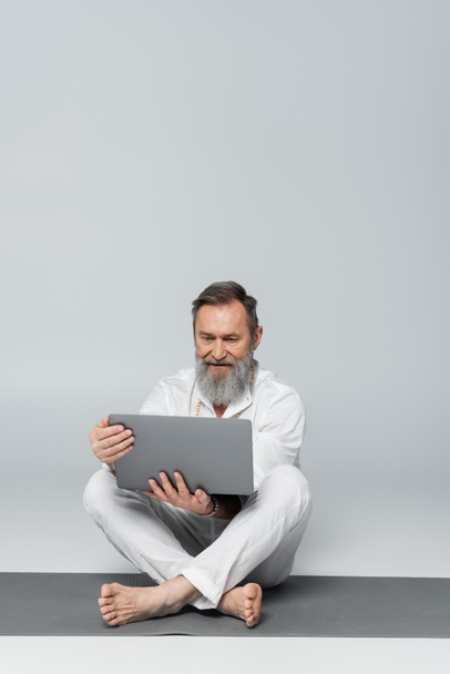 seniorský trenér jógy s laptopem sedícím na podložce jógy v jednoduché póze na šedi - Fotografie, Obrázek