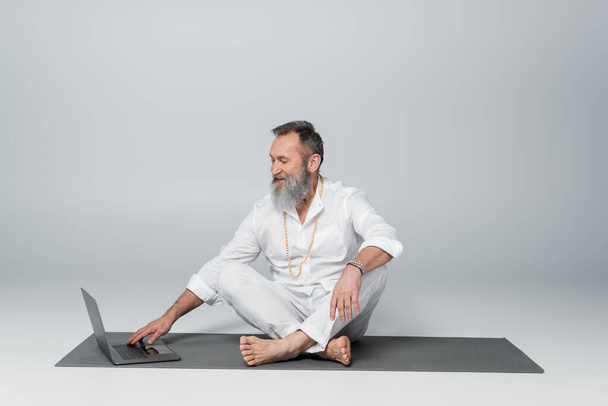 mentor espiritual sonriente sentado en la esterilla de yoga en pose fácil cerca del ordenador portátil en gris - Foto, Imagen
