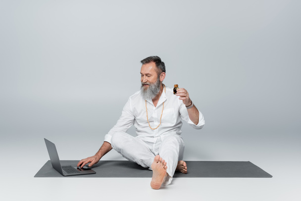 Mezítláb gyógyító guru ül a laptop mellett és egy üveg illatos olajat tart a szürkén. - Fotó, kép