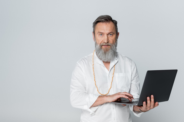 zelfverzekerde spirituele mentor die laptop vasthoudt en kijkt naar camera geïsoleerd op grijs - Foto, afbeelding