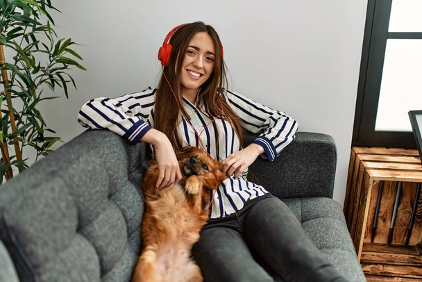 Young hispanic woman listening to music sitting on sofa with dog at home - Fotoğraf, Görsel