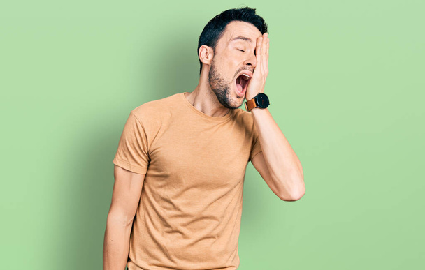 Ισπανόφωνος άντρας με γενειάδα που φοράει casual μπλουζάκι χασμουριέται κουρασμένος καλύπτοντας το μισό πρόσωπο, το μάτι και το στόμα με το χέρι. το πρόσωπο πονάει από τον πόνο.  - Φωτογραφία, εικόνα