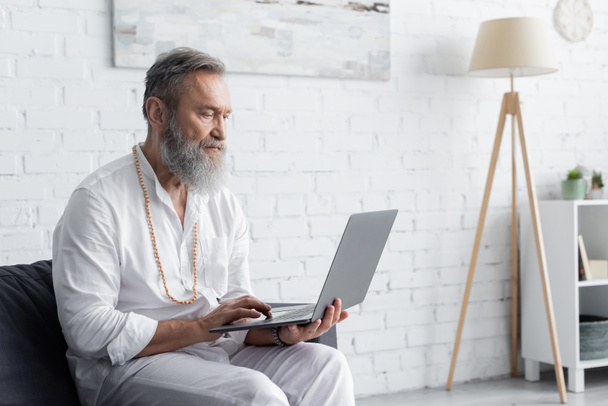 bearded spiritual coach in white clothes sitting on sofa and using laptop - Φωτογραφία, εικόνα