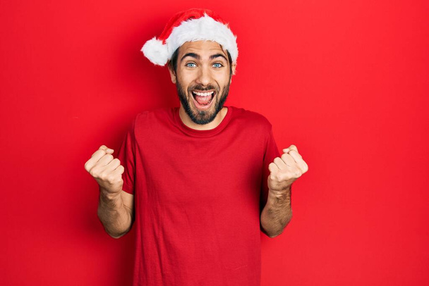 Young hispanic man wearing christmas hat celebrating surprised and amazed for success with arms raised and open eyes. winner concept.  - Foto, imagen