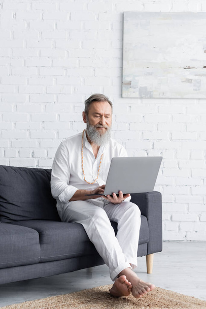 barbuto maestro guru in abiti bianchi seduto con computer portatile sul divano a casa - Foto, immagini