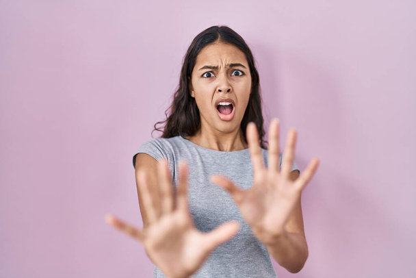 Молодая бразилька в обычной футболке на розовом фоне, напуганная и испуганная выражением страха, прекращает жесты руками, крича в шоке. концепция паники.  - Фото, изображение