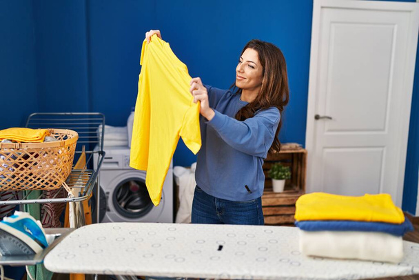 Young hispanic woman smiling confident holding clothes at laundry room - Photo, Image