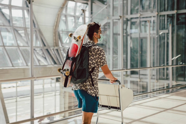 Νεαρός με μάσκα προσώπου που περπατάει στο αεροδρόμιο με σκέιτμπορντ στην πλάτη του - Φωτογραφία, εικόνα