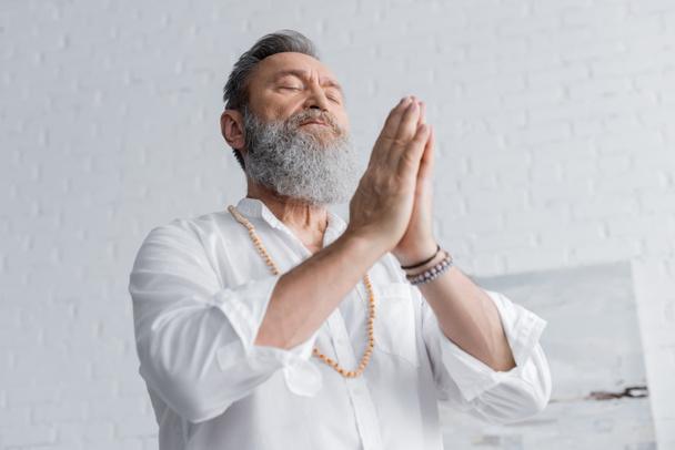 low angle view of bearded guru man meditating with closed eyes and praying hands - Foto, immagini