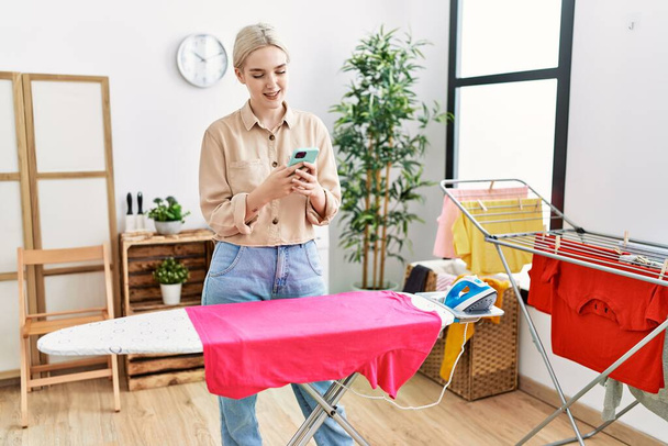 Young caucasian woman using smartphone ironing clothes at laundry room - Valokuva, kuva