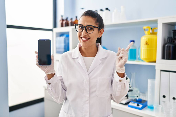 Young hispanic woman working at scientist laboratory with smartphone smiling happy pointing with hand and finger to the side  - Foto, Imagem