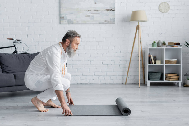 Seitenansicht eines älteren Mannes in weißer Kleidung, der zu Hause eine Yogamatte ausrollt - Foto, Bild