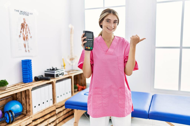 Mladá fyzioterapeutka pracující na klinice pro zotavení bolesti držící datafon ukazující palcem nahoru na stranu usmívající se spokojeně s otevřenými ústy  - Fotografie, Obrázek