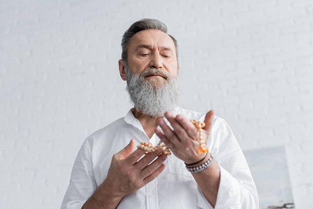 low angle view of bearded guru man meditating with mala beads at home - Foto, immagini