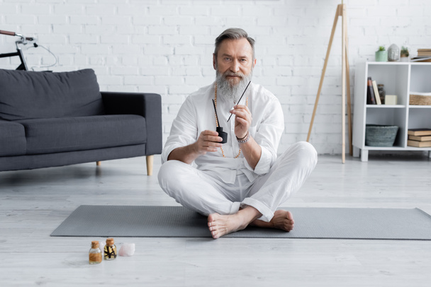 vousatý hojící guru sedí v pohodlné póze s aromatickými tyčinkami v blízkosti lahví s ochucenými oleji - Fotografie, Obrázek