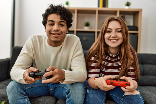 Młoda para uśmiechnięta szczęśliwa grając w gry wideo siedząc na kanapie w domu. - Zdjęcie, obraz