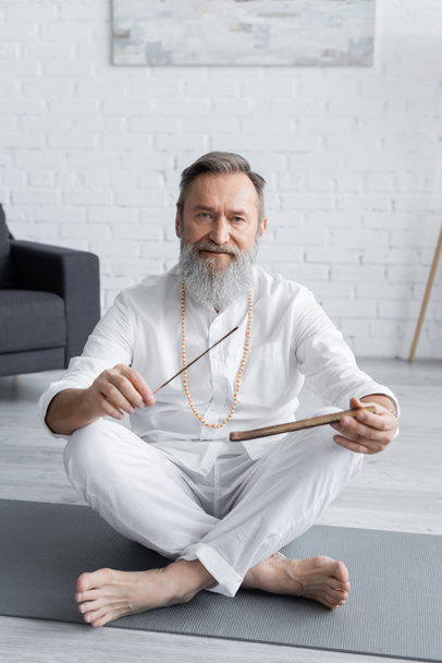 bearded yoga teacher holding aroma sticks while sitting in easy pose and looking at camera - Photo, Image