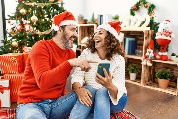 Ein hispanisches Paar mittleren Alters lächelt glücklich mit Weihnachtsmütze. Zuhause mit Smartphone auf dem Boden sitzen. - Foto, Bild
