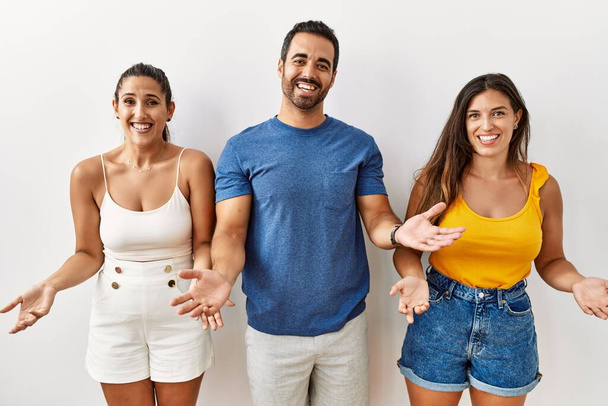 Группа молодых латиноамериканцев, стоящих на изолированном фоне, радостно улыбающихся с распростертыми объятиями, как дружеские приветствия, позитивные и уверенные приветствия  - Фото, изображение