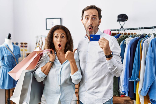Hispánský pár středního věku držící nákupní tašky a kreditní kartu překvapeně ukazuje ukazováčkem na stranu, otevřená ústa užaslý výraz.  - Fotografie, Obrázek