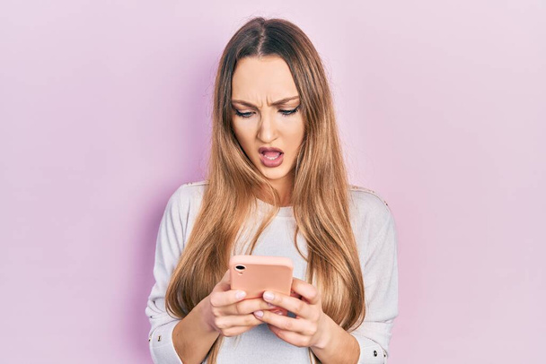 Young blonde girl using smartphone typing message in shock face, looking skeptical and sarcastic, surprised with open mouth  - Fotoğraf, Görsel