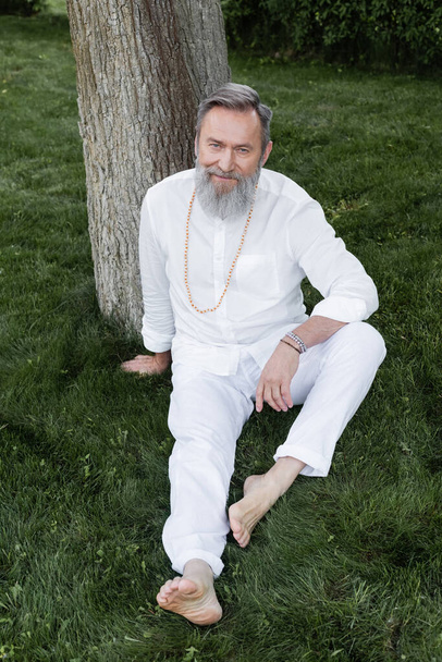 plná délka vousaté jógy trenér sedí na zeleném trávníku a usmívá se na kameru - Fotografie, Obrázek