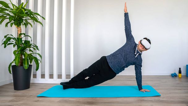 Чоловік сидить у спортивному одязі на килимку для йоги у вітальні в сонячний день. Спортсмен дивиться 360 бачення віртуальної реальності для спортивної рутини. Насолоджуючись кібер-досвідом у концепції реальності моделювання vr
 - Фото, зображення