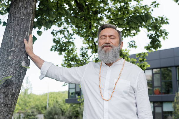 man met baard in wit shirt en kralen mediteren met gesloten ogen in de buurt van boom - Foto, afbeelding