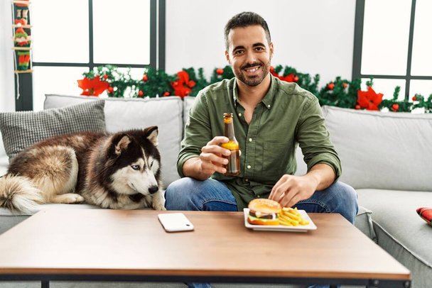 Young hispanic man eating hamburger sitting on sofa with dog by christmas decor at home - Foto, immagini