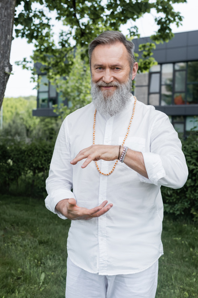 glimlachende goeroe man in wit shirt toont energie gebaar tijdens het mediteren buiten - Foto, afbeelding