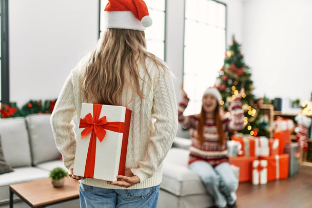 Mujer pareja sorpresa con regalo en espalda sentado por árbol de Navidad en casa - Foto, Imagen
