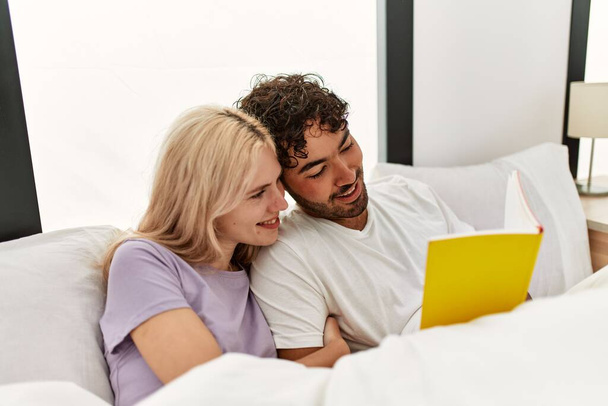 Young beautiful couple reading book lying in bed at home. - Фото, изображение