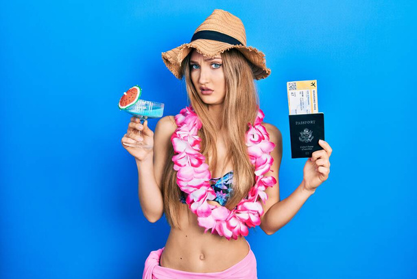 Young blonde girl wearing bikini holding cocktail and passport clueless and confused expression. doubt concept.  - 写真・画像