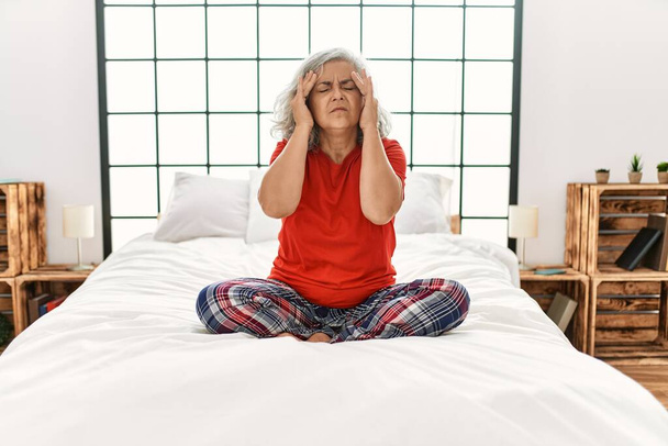 Kobieta w średnim wieku z siwymi włosami siedząca na łóżku w domu z ręką na głowie na ból głowy, ponieważ stres. migrena.  - Zdjęcie, obraz