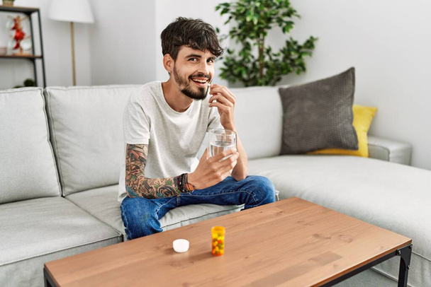 Young hispanic man taking pills sitting on the sofa at home - Photo, image